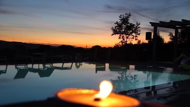 Área tranquila de piscina al aire libre al anochecer, con una vela encendida, sillas de descanso y una pérgola.