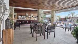 An outdoor dining area with wooden tables and chairs, a bar in the background, and a pool area visible through large windows. - Holiday Inn Cairo Maadi (Cairo)