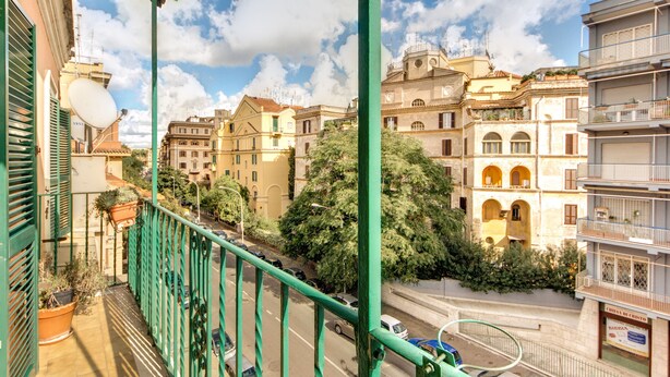 A balcony with a view of a street lined with buildings, a clear sky, and some greenery. - Attico Sannio 1 (Rome)