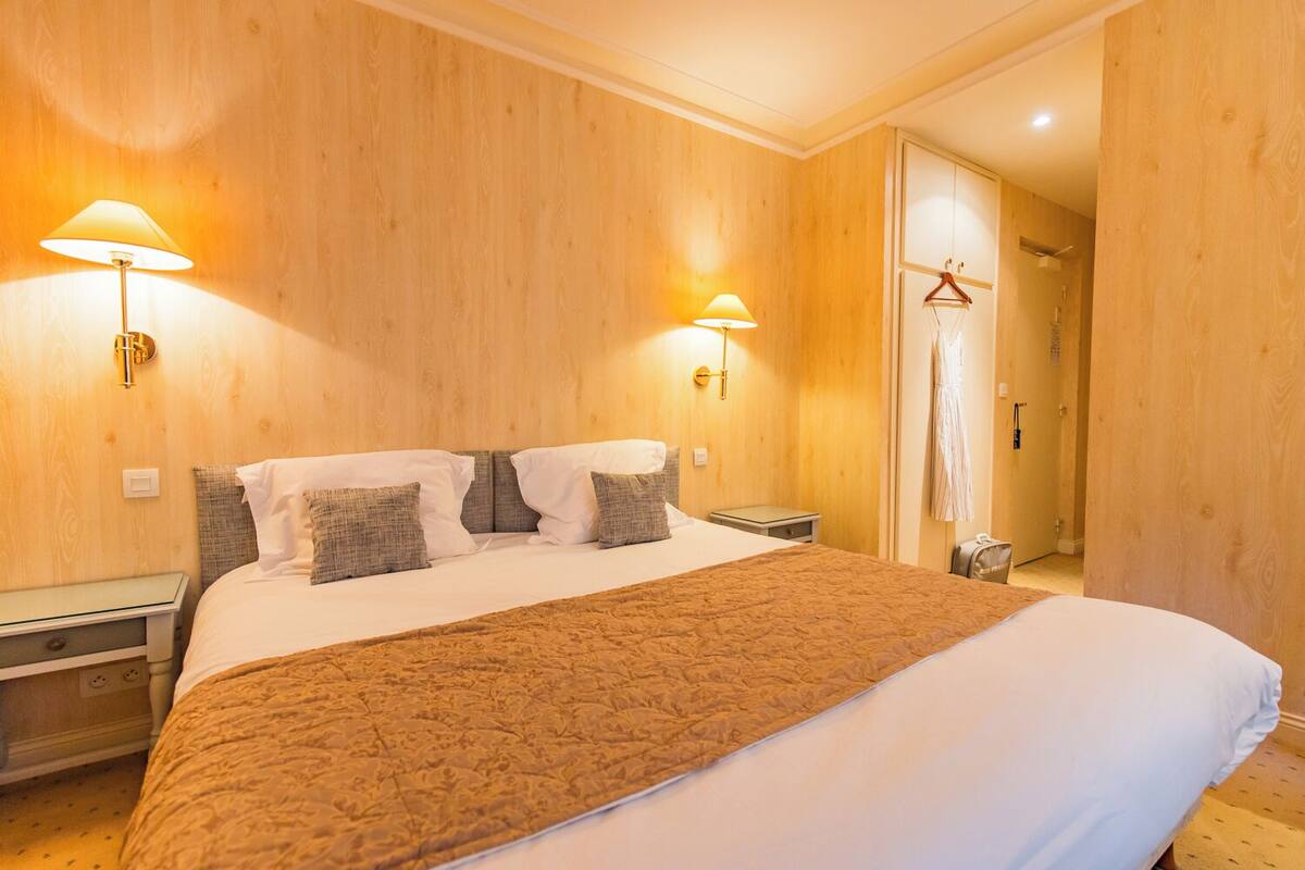 A hotel room with a large bed, two bedside tables, a wooden headboard, and two wall-mounted lamps.