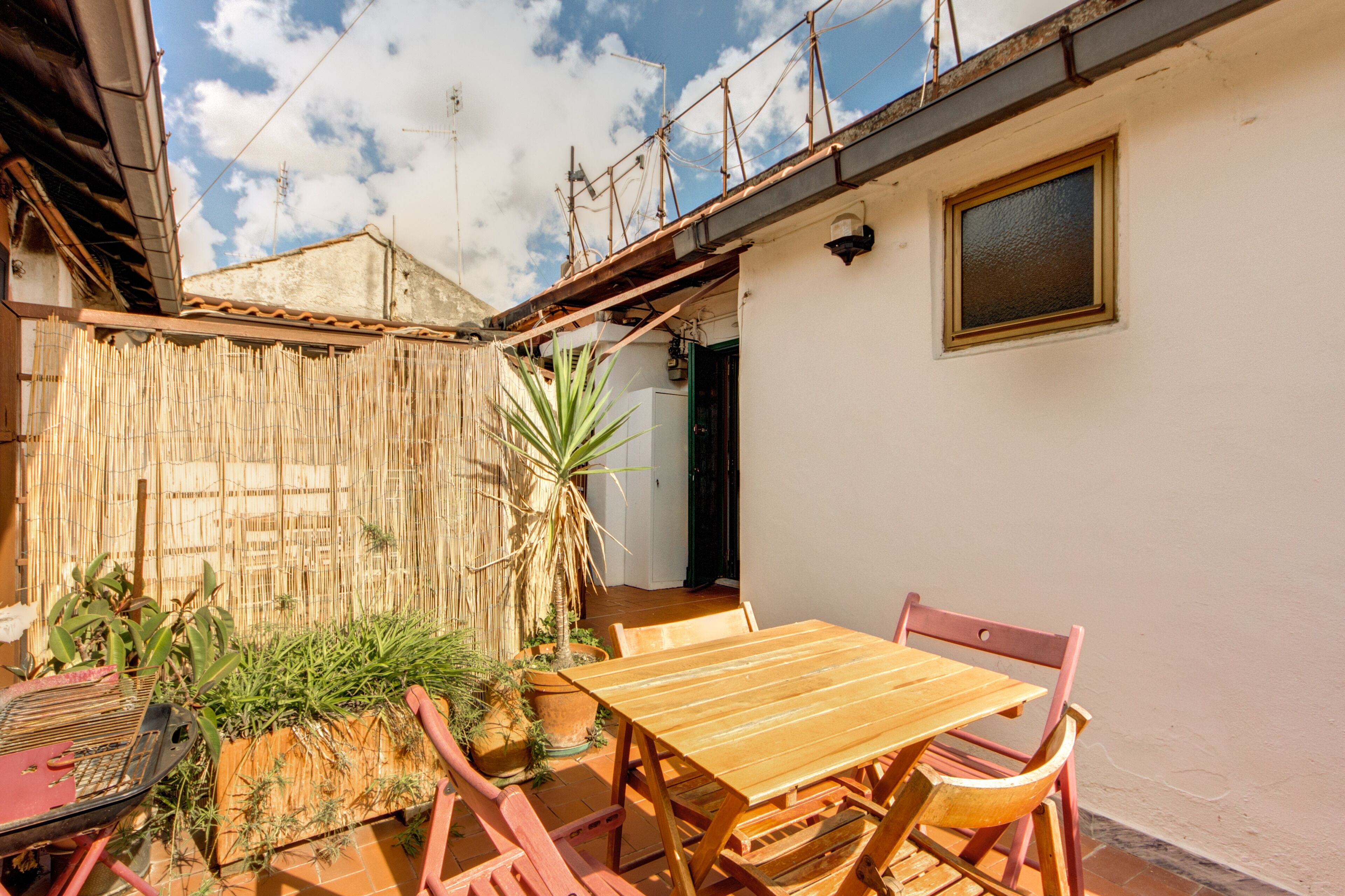 Una mesa y sillas de madera en un patio con cerca de bambú, plantas en macetas y vista a un edificio con ventana y puerta.
