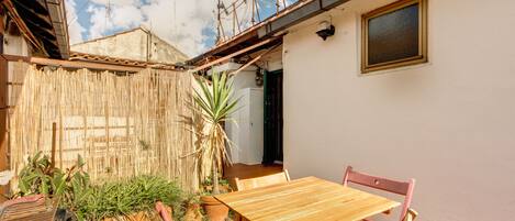 Una mesa y sillas de madera en un patio con cerca de bambú, plantas en macetas y vista a un edificio con ventana y puerta.