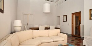 Una sala de estar con un sofá color crema, una mesa de comedor con sillas, una lámpara y una puerta que da a otra habitación.