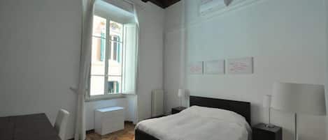 Quarto com cama, mesa de cabeceira, abajur, cômoda e janela com cortinas.