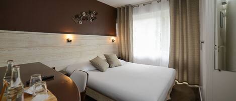 Habitación de hotel impecable con cama, mesita de madera con botella y vasos, una silla y una lámpara de pared.