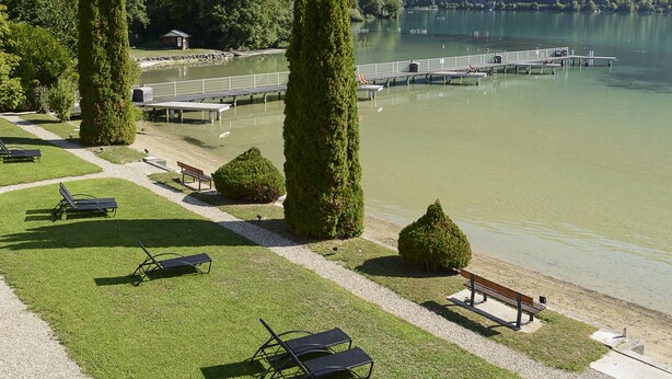 A lakeside resort with grassy areas, lounge chairs, and a dock extending into the water. - Hôtel Restaurant Novalaise Plage (Novalaise)