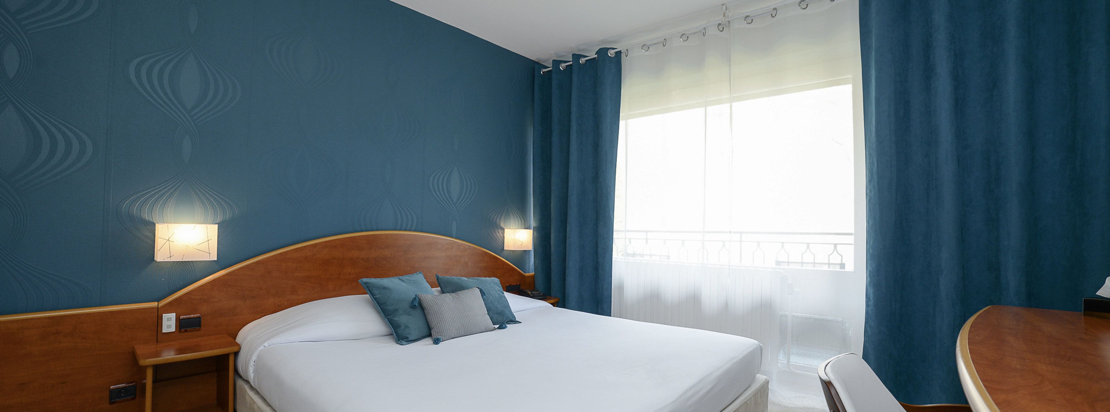 Quarto de hotel com uma cama grande, duas mesas de cabeceira, uma escrivaninha e uma janela com cortinas.