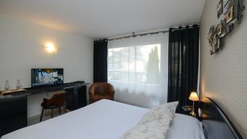 A hotel room with a bed, a desk with a computer, a chair, a window with curtains, and a wall-mounted light fixture.
