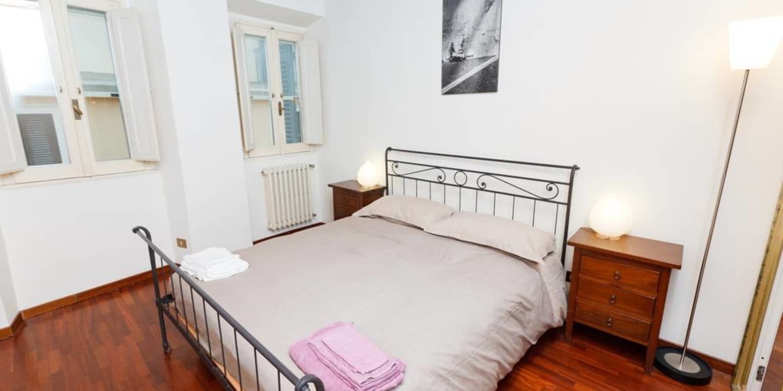 Une chambre à coucher comprenant un lit, une table de chevet, une commode, un radiateur et une fenêtre avec des volets.