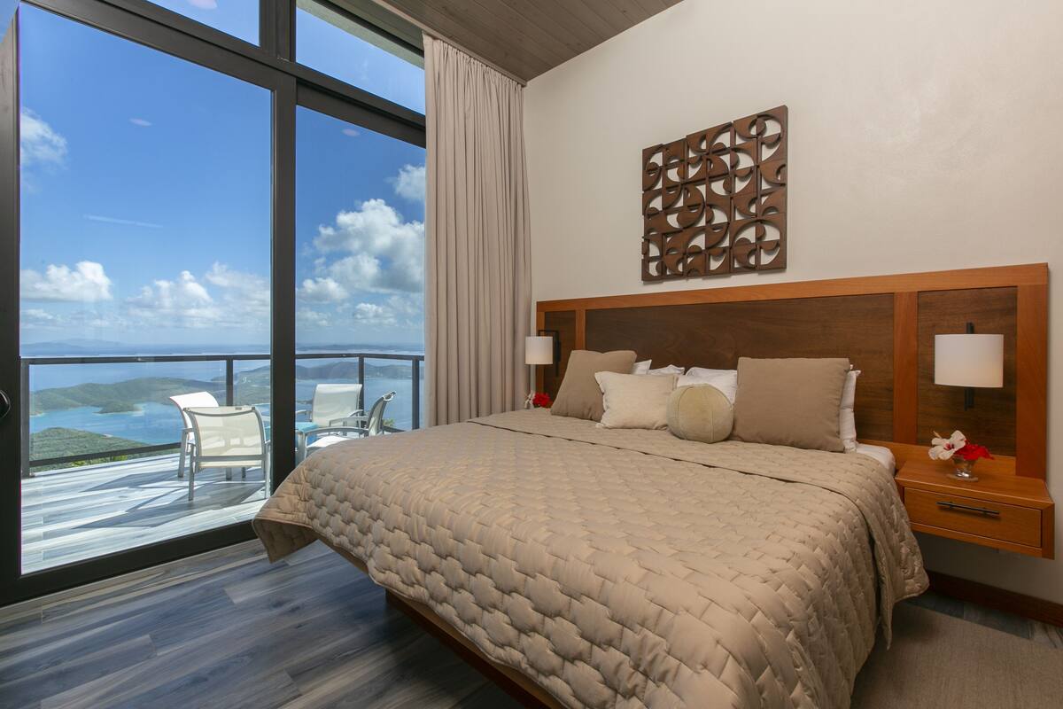 A bedroom with a large bed, a wooden headboard, two bedside tables with lamps, a decorative wall piece, and a view of the sea through floor-to-ceiling windows.