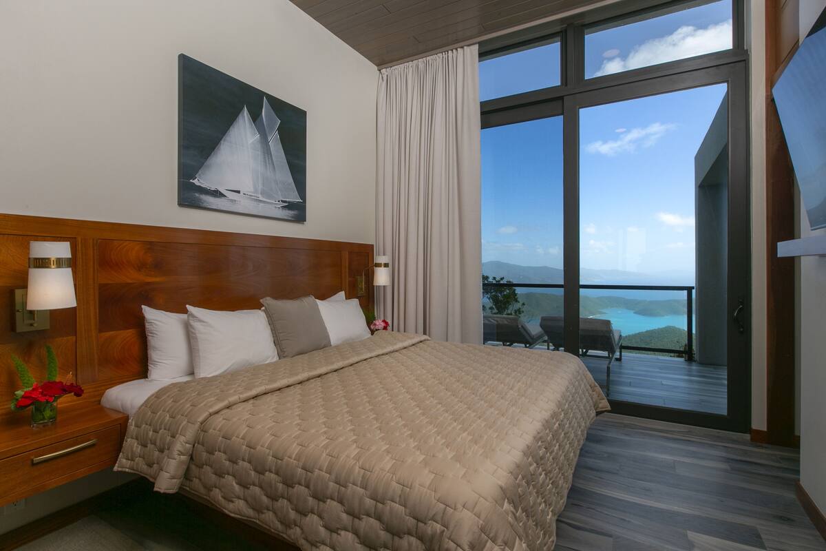 A hotel room with a large bed, wooden headboard, a painting of a sailboat, a nightstand with a lamp, and a view of the ocean through large windows.