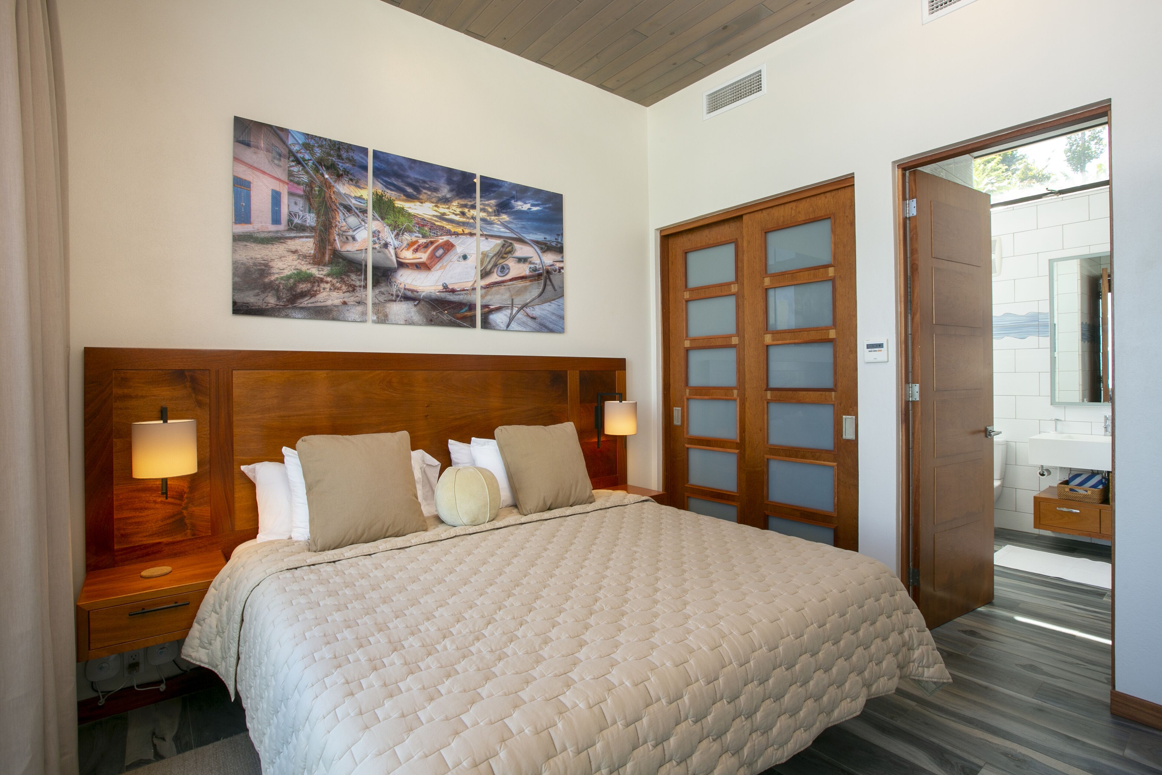 A hotel room with a large bed, wooden headboard, two bedside tables with lamps, a wooden door, and a wall-mounted painting.