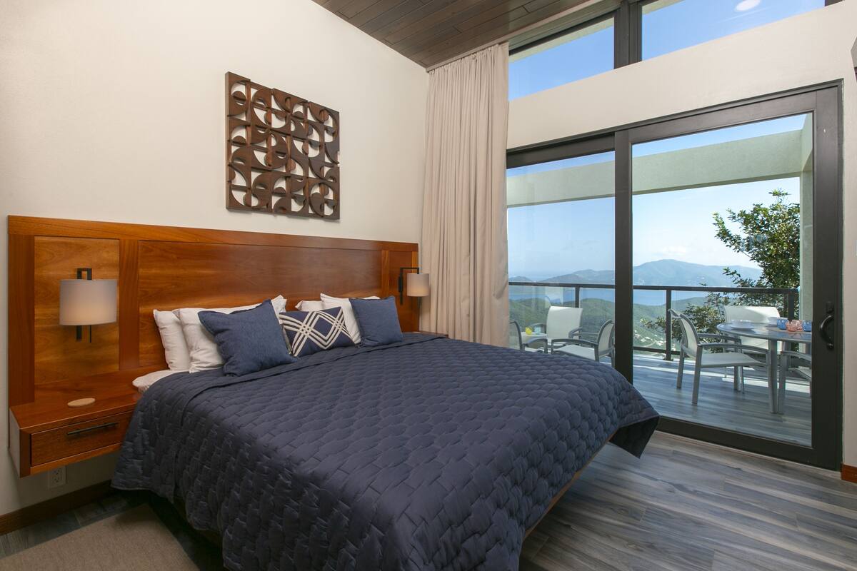 A modern hotel room with a large bed, a wooden headboard, a decorative wall piece, and a balcony with a view of the mountains.