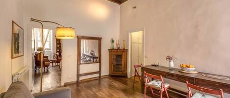 A spacious living room with a wooden floor, a sofa, a dining area with a wooden table and chairs, and a large mirror.