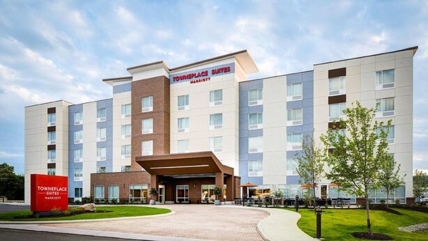Un hotel TownePlace Suites Marriott con diseño moderno, que presenta una entrada central, varios pisos y un área exterior ajardinada con árboles y un patio.