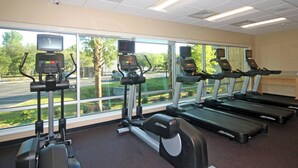 Un gimnasio con cintas de correr, elípticas y bicicletas estáticas, amplias ventanas que dan a un estacionamiento y zonas verdes.