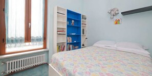 Une chambre à coucher avec un lit, un radiateur, une fenêtre avec des rideaux, une bibliothèque et une lampe fixée au mur.