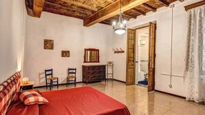 A bedroom with a wooden ceiling, a bed with a red bedspread, two chairs, a dresser, and a door leading to a bathroom. - Campo de Fiori 3 bedroom apartment (Rome)