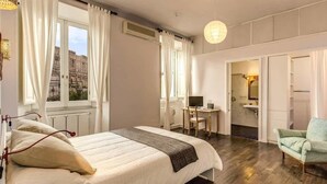 A hotel room with a bed, a desk, a chair, and a view of a historic building through the window. - Colosseo view Suite (Rome)