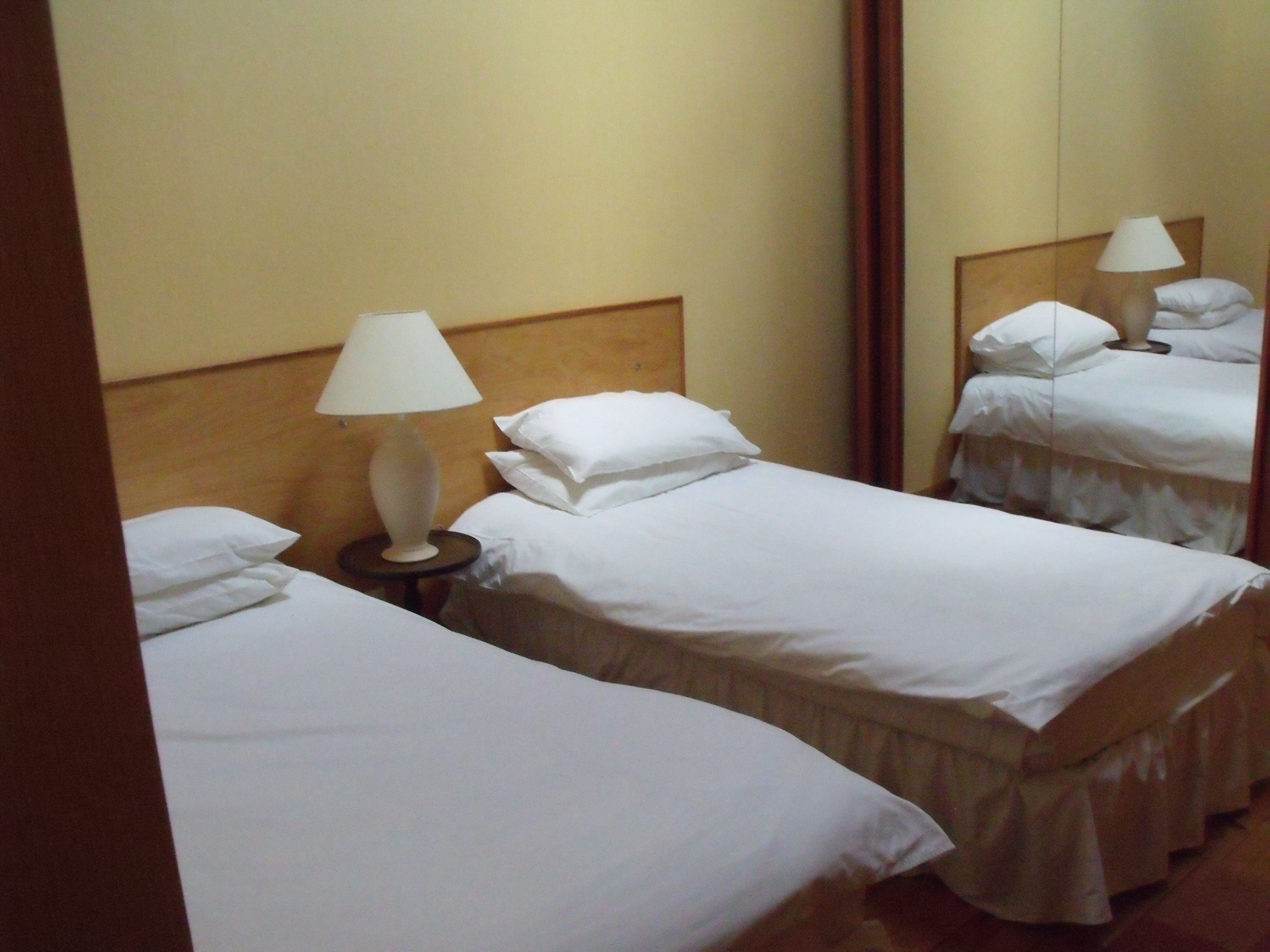 Habitación de hotel con dos camas individuales, una mesita de noche con lámpara y un espejo grande que refleja la habitación.