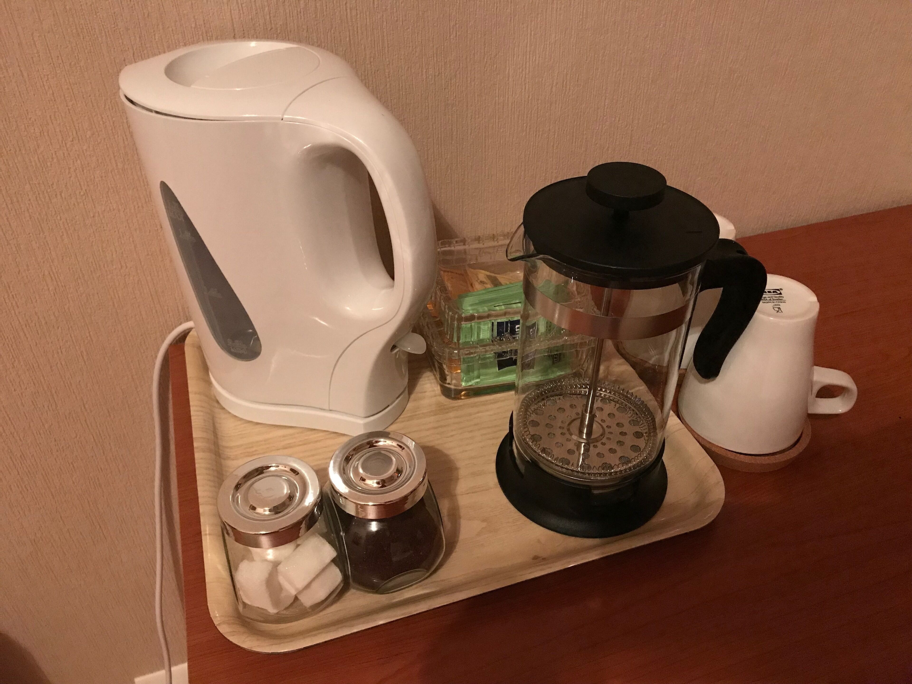 Una encimera de cocina con un hervidor eléctrico blanco, una cafetera de vidrio, dos tarros de vidrio con tapas y una taza blanca.