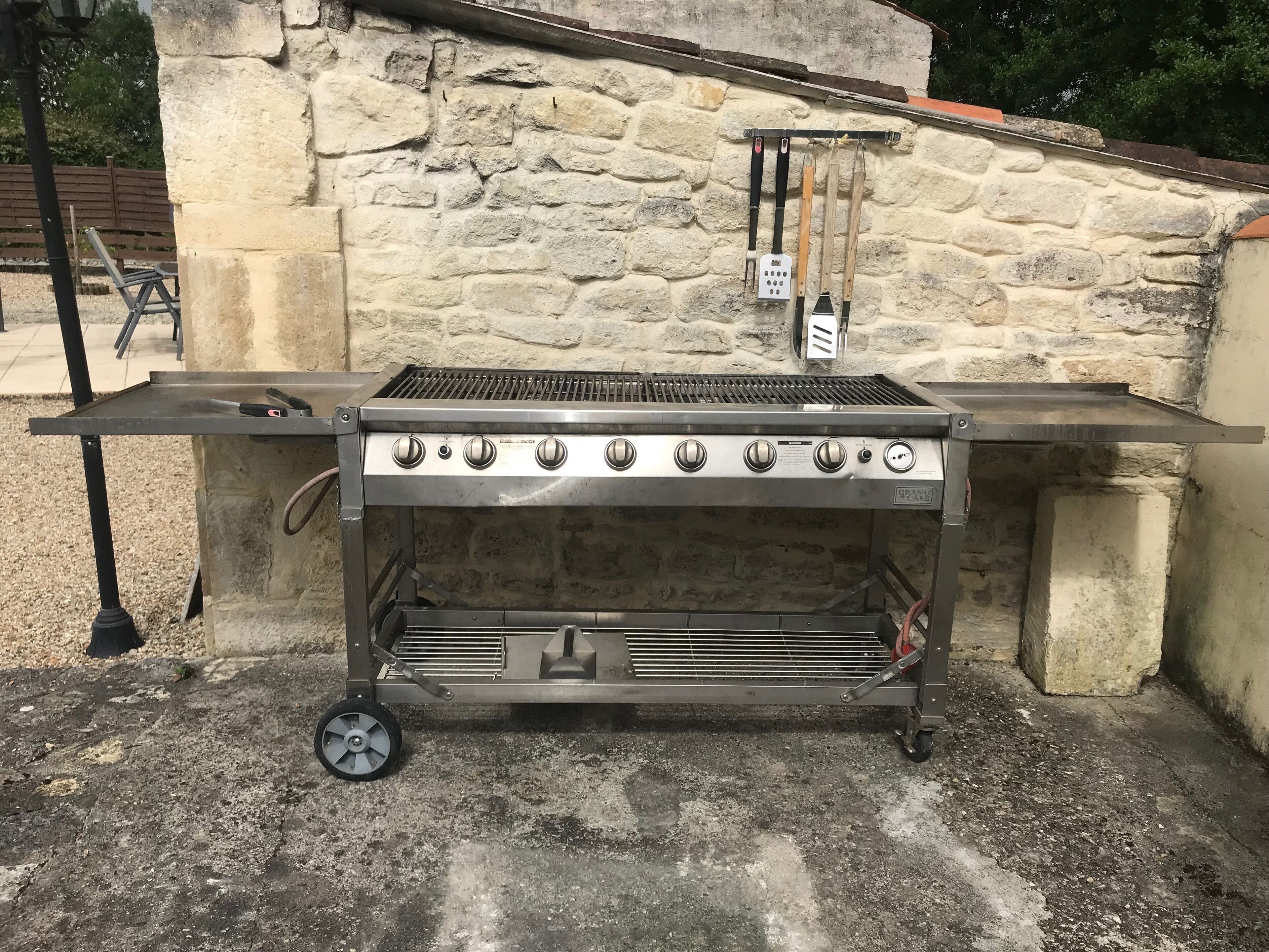 A portable stainless steel grill with five burners, wheels, and a side shelf, set against a stone wall.