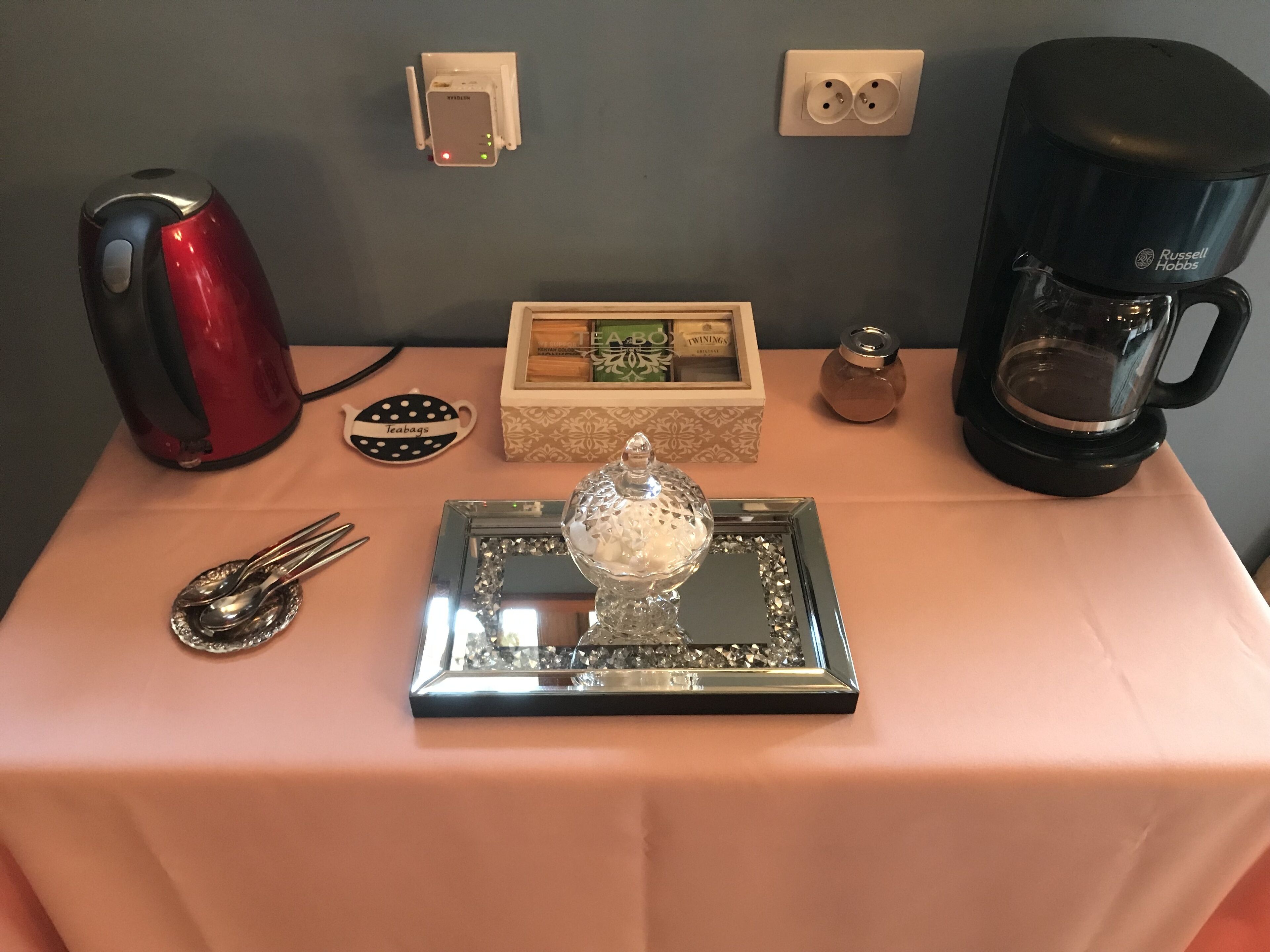 Una mesa con un calentador de agua, tazas de té, una tetera, una bandeja, infusiones envasadas y una cafetera.
