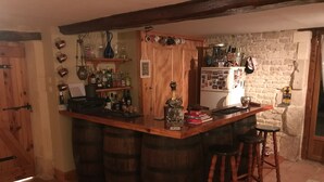 A rustic bar area with wooden barrels as stools, a wooden counter, and various bottles and glasses. - Chambre d'hôtes Les Cygnes Noirs (Aujac)