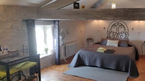 Une chambre confortable avec un plancher en bois, un lit recouvert d’une courtepointe grise, une petite table avec une chaise et une fenêtre avec des rideaux.