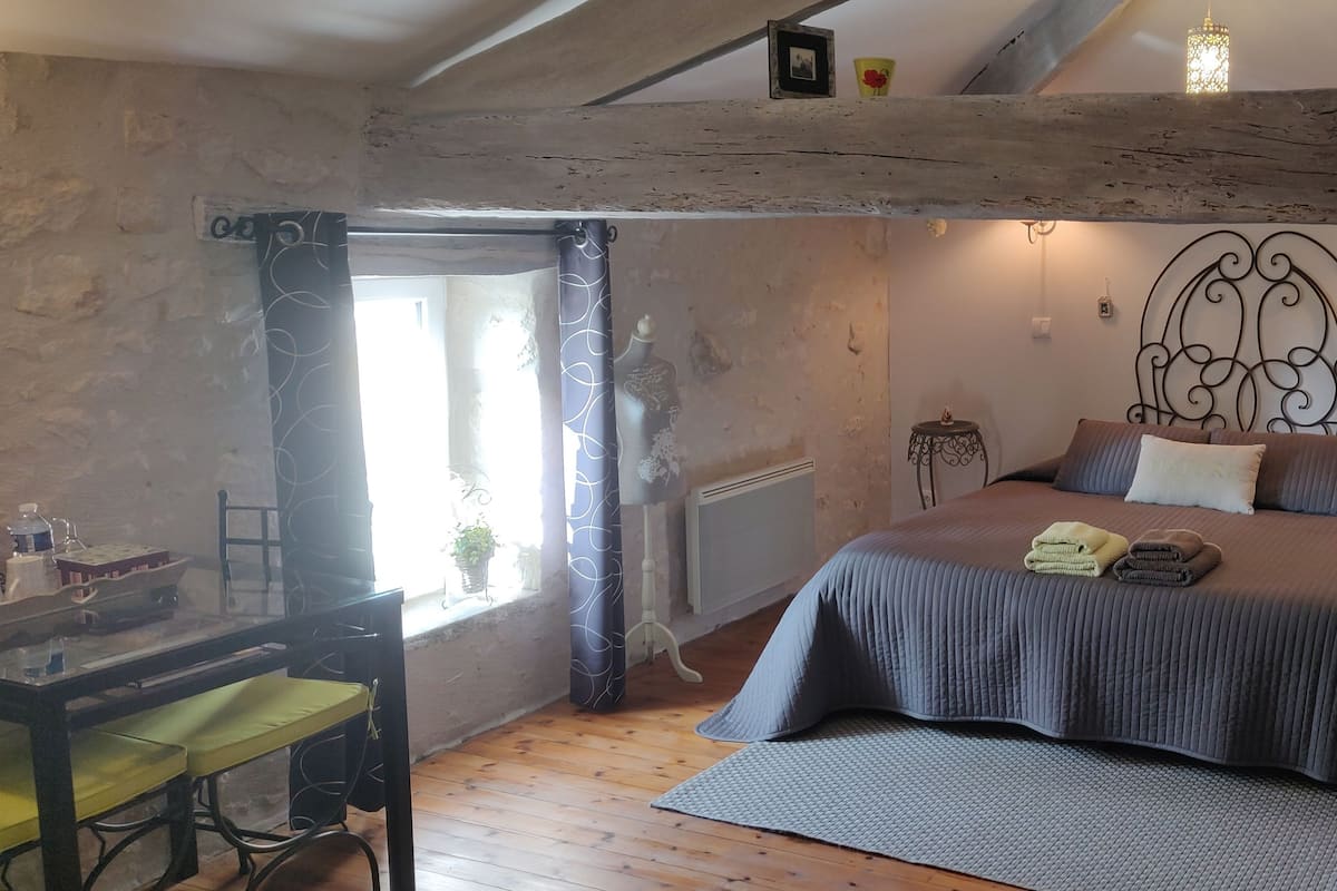 Un dormitorio acogedor con piso de madera, una cama con cubrecama gris, una mesita con una silla y una ventana con cortinas.