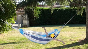 Un hamac est tendu entre deux arbres dans une cour verdoyante, avec un mur de pierres et un toit en tuiles en arrière-plan.