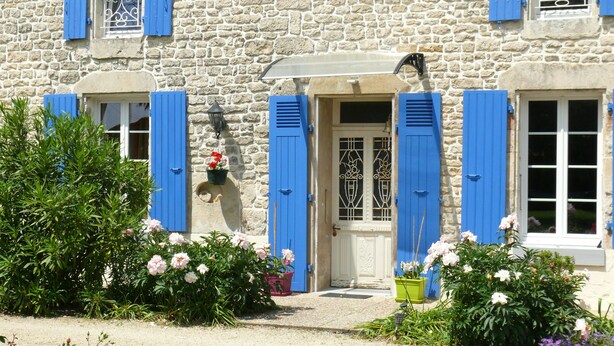 Un bâtiment en pierre avec des volets bleus, une porte blanche et un panier à fleurs suspendu.