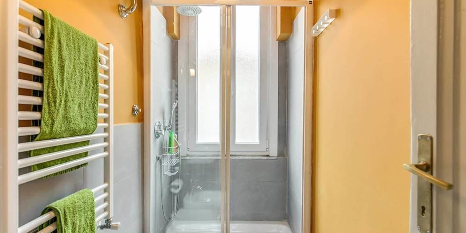 A bathroom with a glass-enclosed shower, a towel rack with green towels, and a window.
