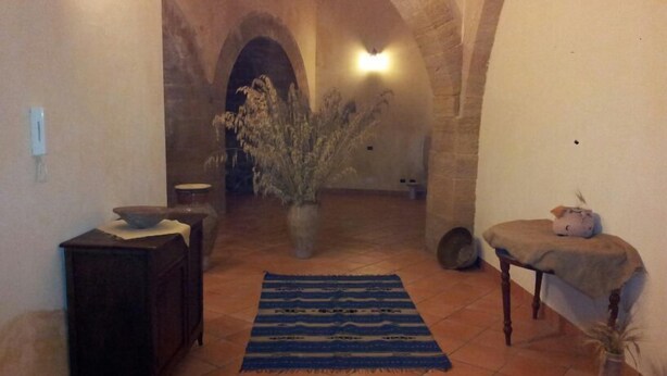 A hallway with a rustic stone archway, a potted plant, a blue patterned rug, a small table with a cat tree, and a dark wooden cabinet. - B&B Portagibli (Salemi)