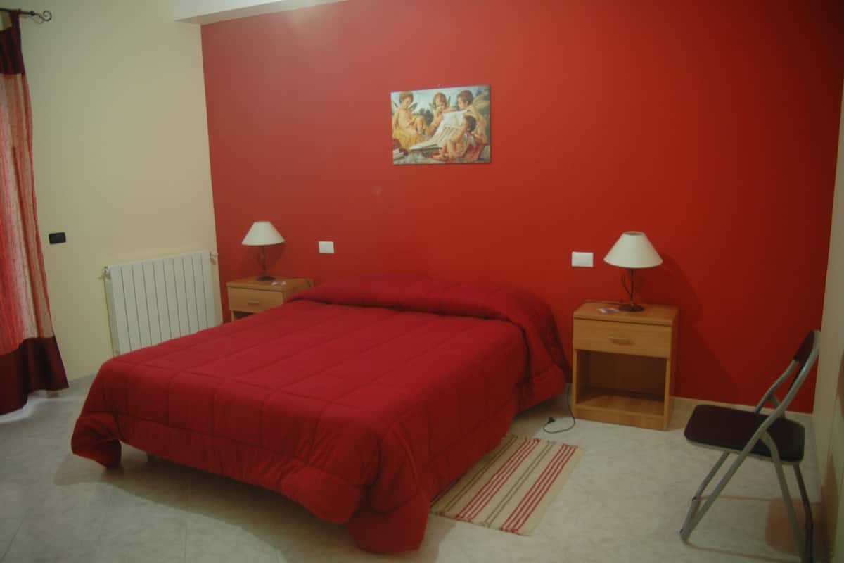 A bedroom with a red bedspread, two bedside tables with lamps, a wooden nightstand, a chair, and a painting on the wall.