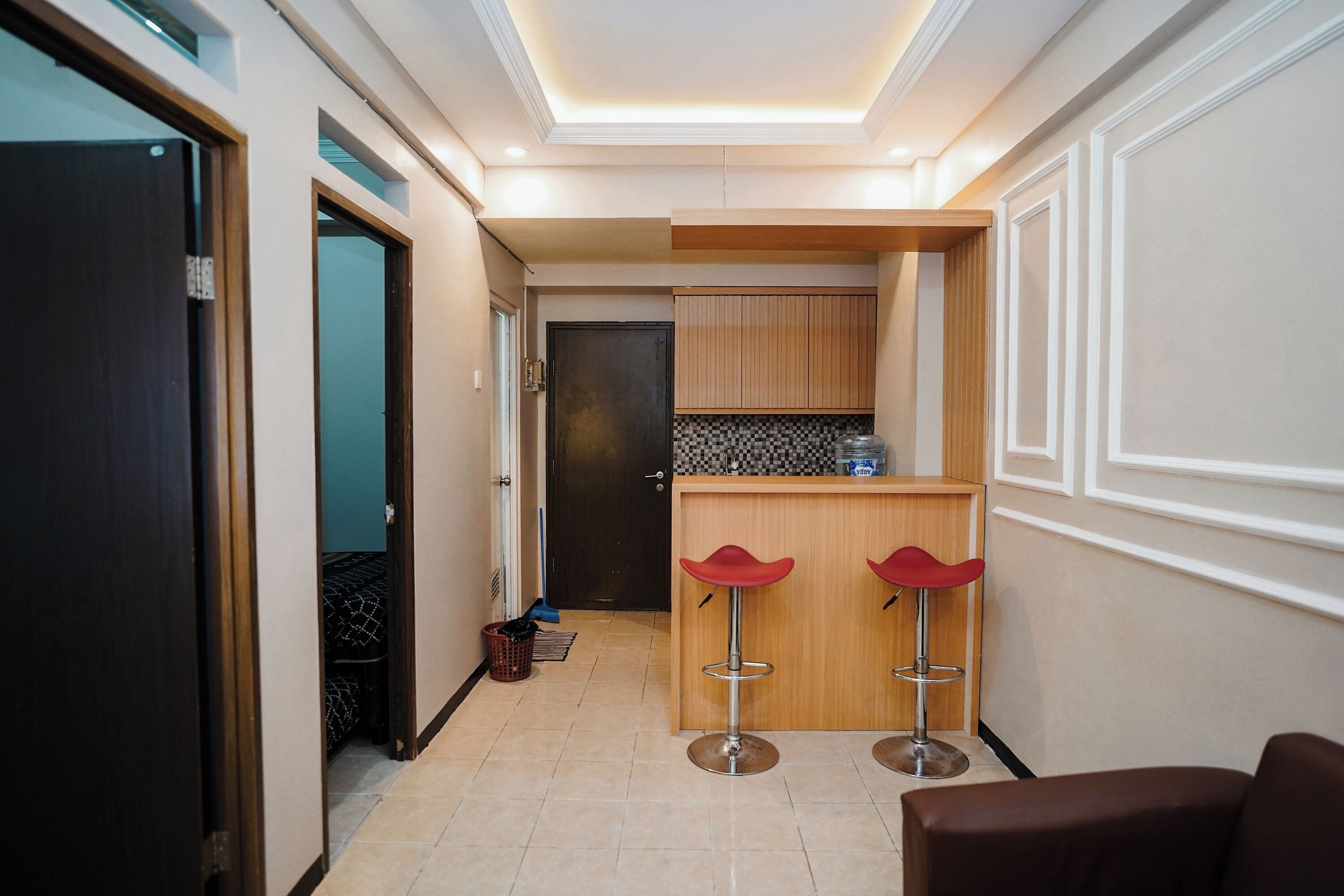A hallway with a kitchen area, two red bar stools, and a brown chair.