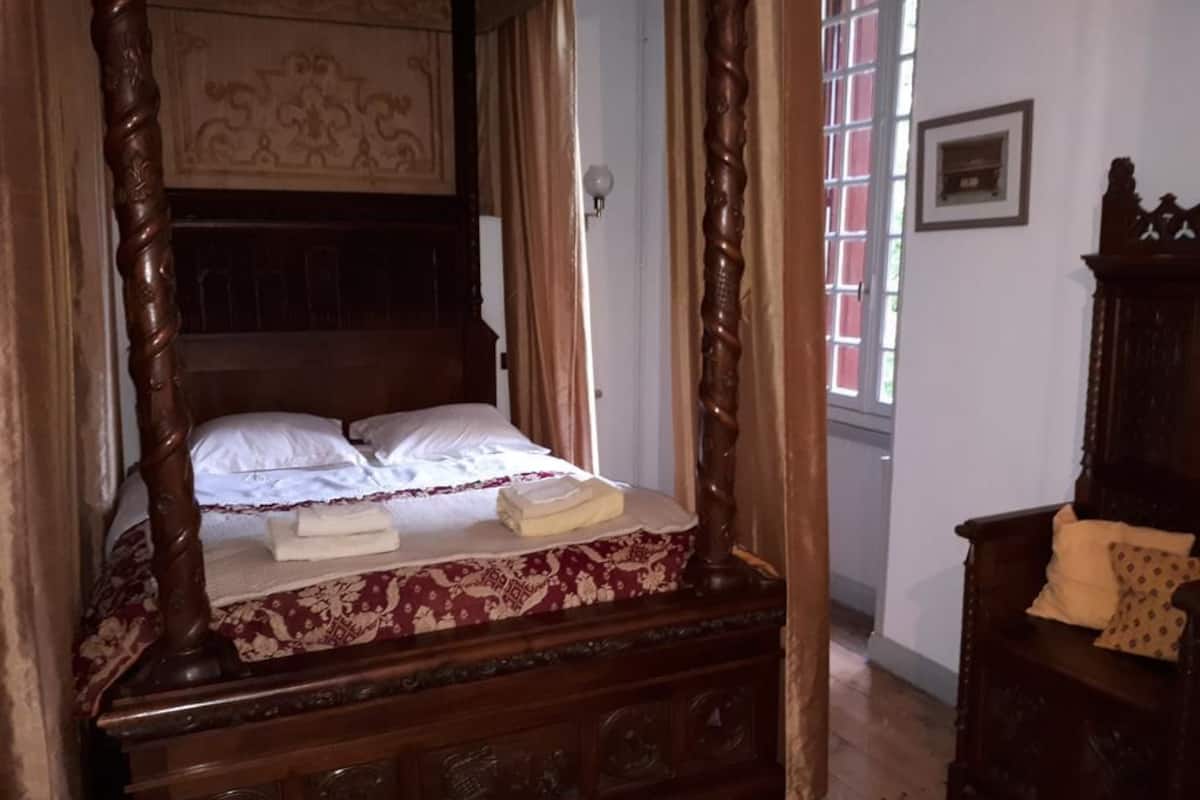 Un lit traditionnel à baldaquin, recouvert d’une courtepointe à motifs floraux et agrémenté de deux serviettes pliées. Une commode en bois avec un miroir se trouve à droite.