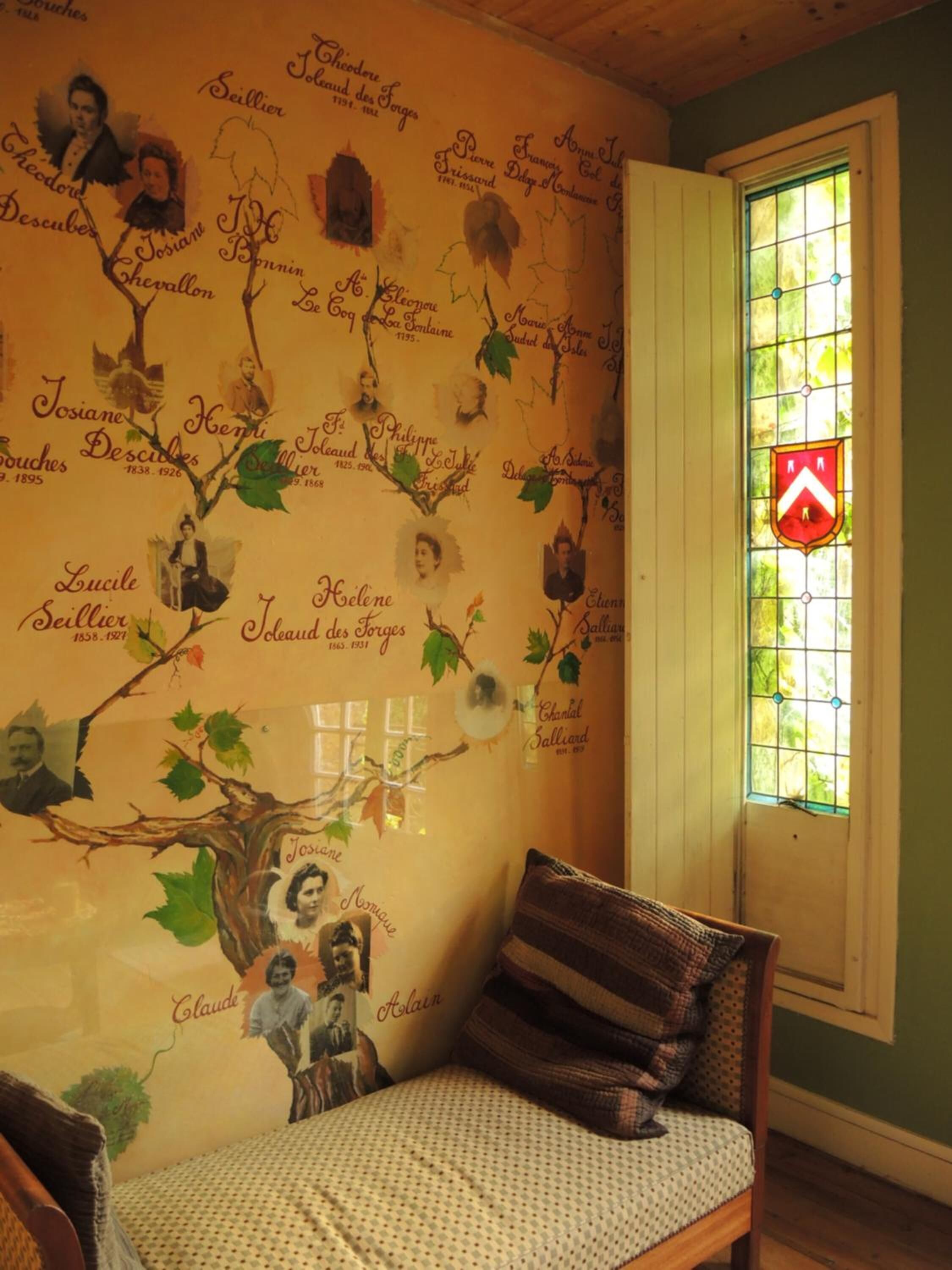 Una habitación con un mural que representa un árbol genealógico, una ventana con un diseño de vitral y un cojín estampado sobre una silla de madera.