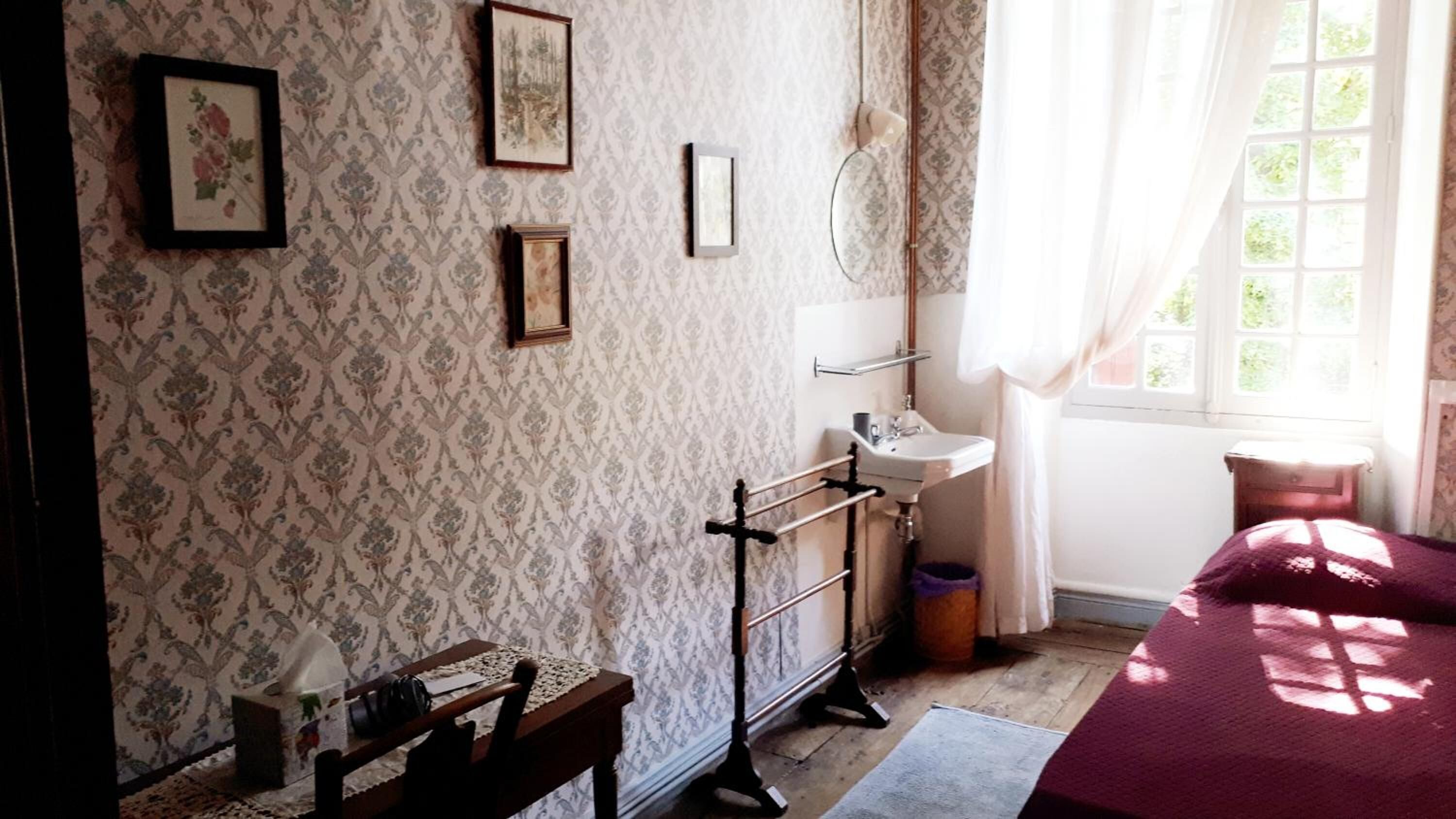 A room with patterned wallpaper, a wooden chair, a small table, a bed with a red blanket, a window with curtains, and a wall-mounted mirror.