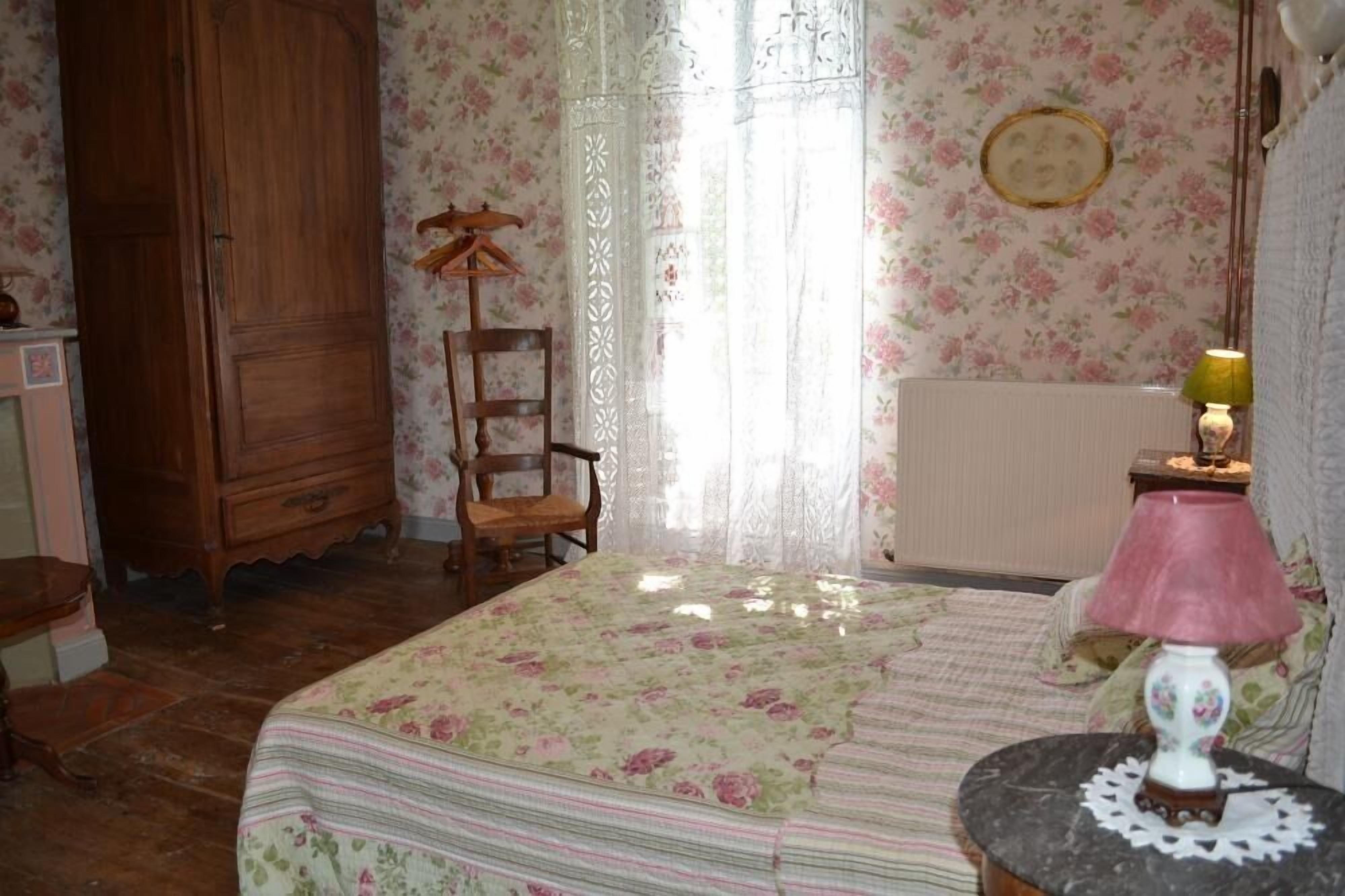 A bedroom with a bed, a wooden wardrobe, a chair, a table with a lamp, and floral wallpaper.