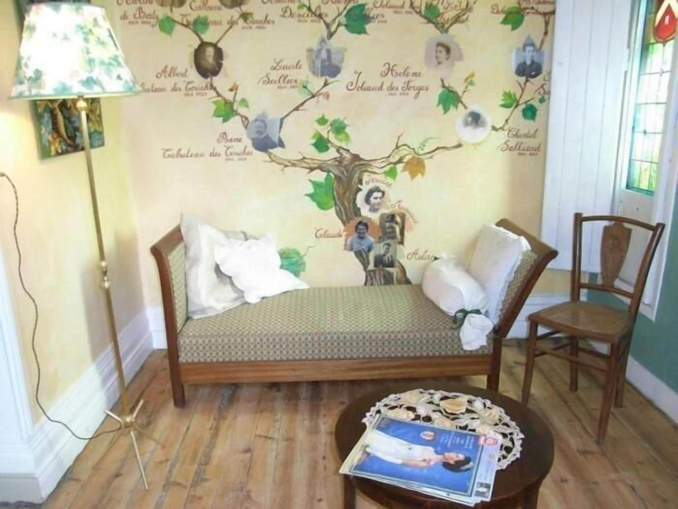 Une pièce chaleureuse avec un canapé en bois, une petite table ronde sur laquelle repose un magazine, et un mur orné d’une fresque représentant un arbre.