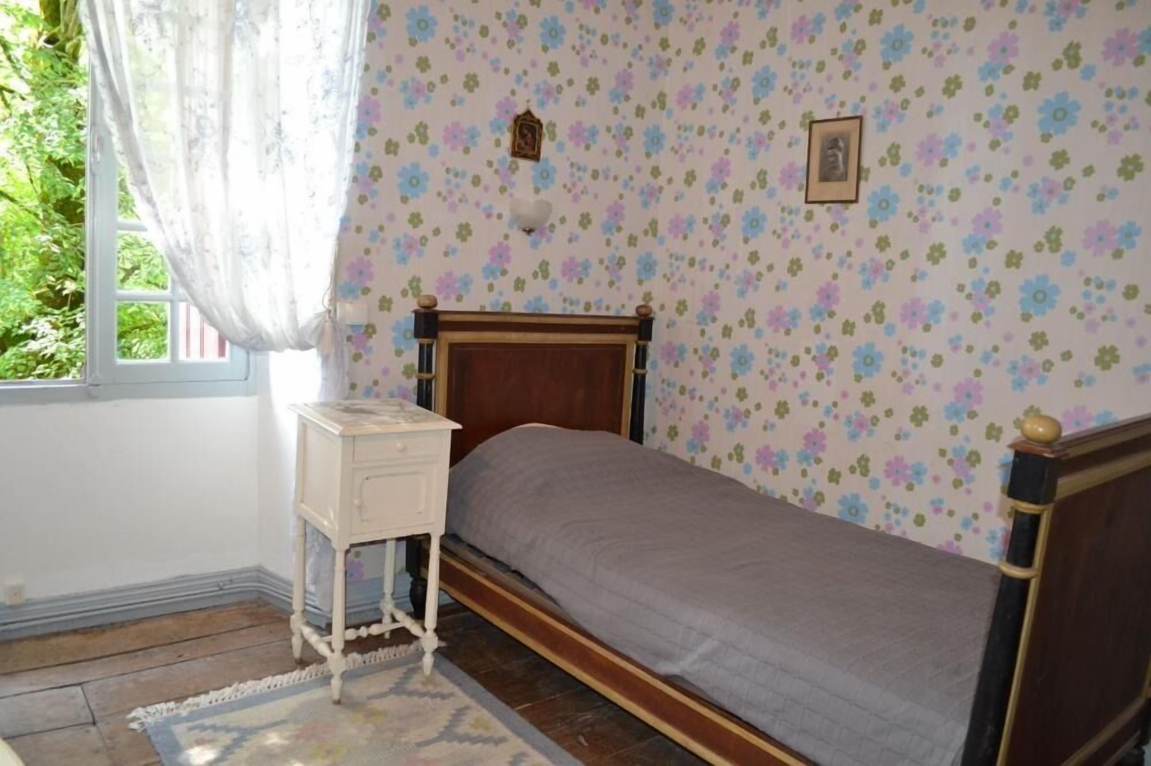 Un dormitorio con una cama de madera, una mesita de noche pequeña, papel tapiz floral y una ventana con cortinas.