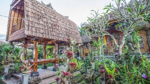 Una casa de madera tradicional con techo de paja, rodeada de exuberante vegetación tropical y un sendero decorativo de piedra.