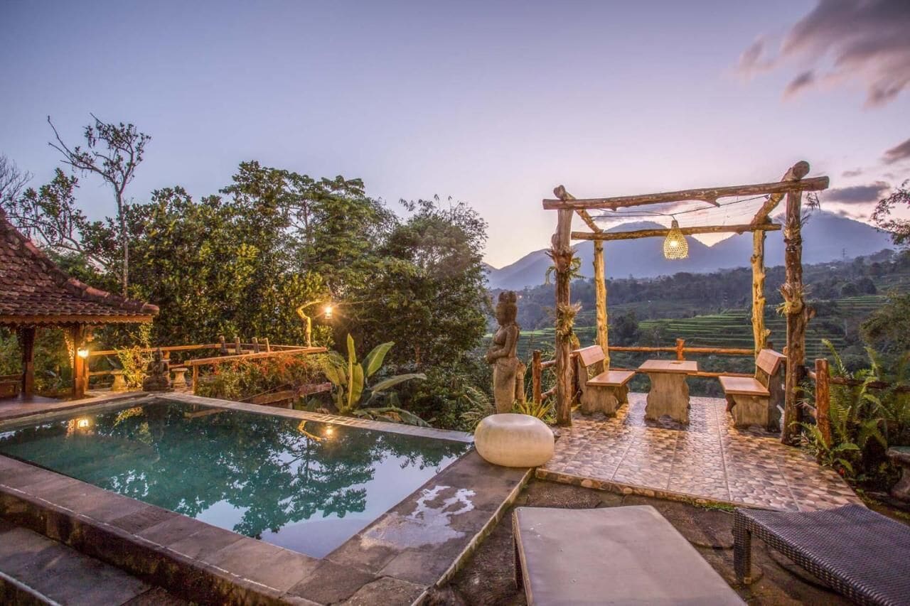 A serene outdoor pool area with a wooden pergola, stone benches, and a view of a mountainous landscape.