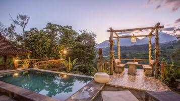 A serene outdoor pool area with a wooden pergola, stone benches, and a view of a mountainous landscape.