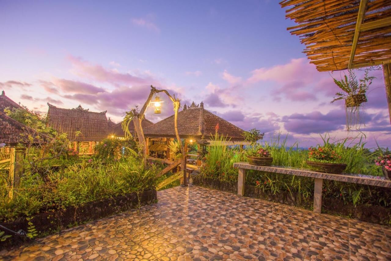 A traditional thatched-roof building with a stone-paved entrance, surrounded by lush greenery and a wooden fence.