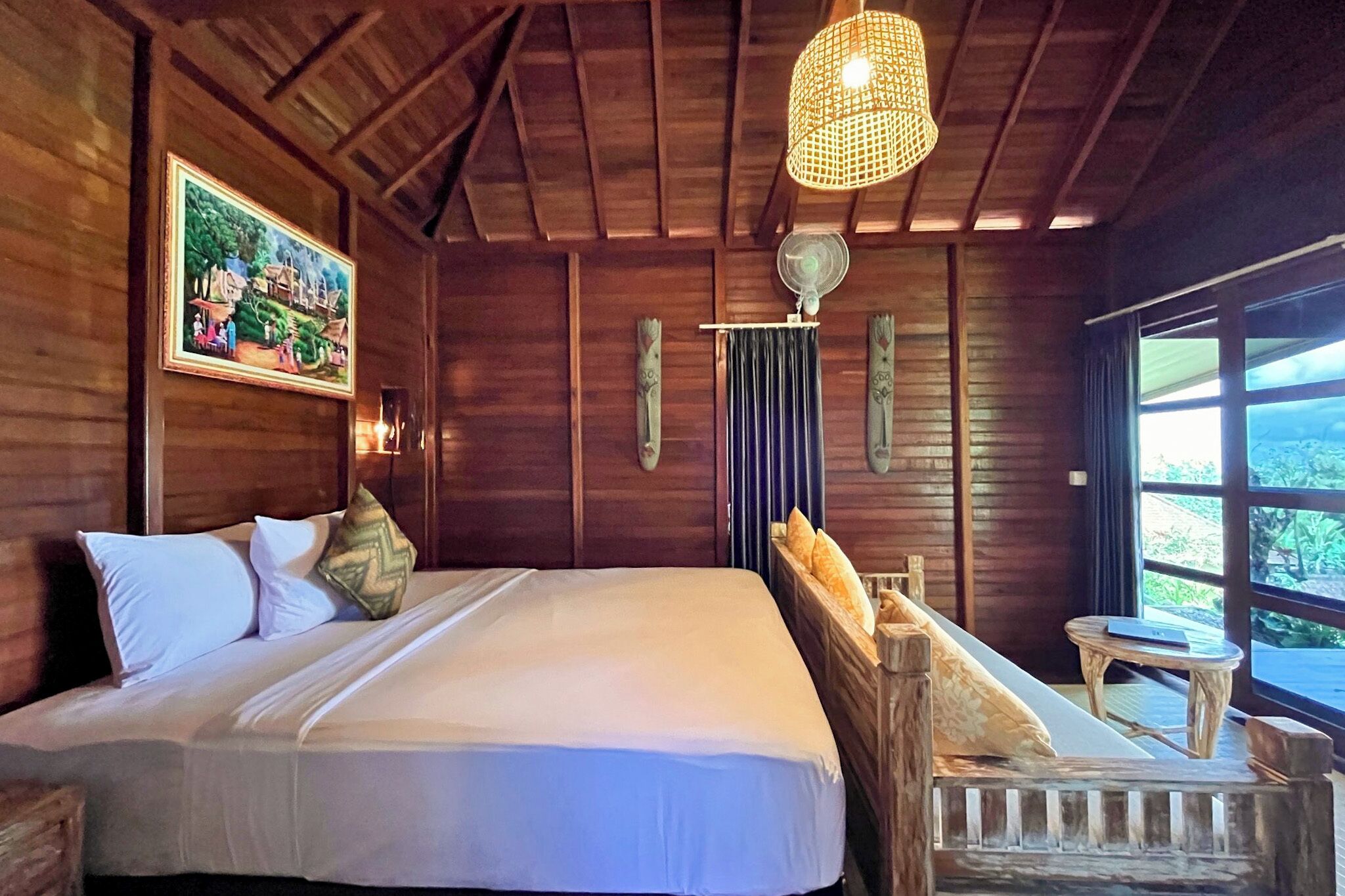 A wooden room with a bed, a small table, and a window with a view of greenery.
