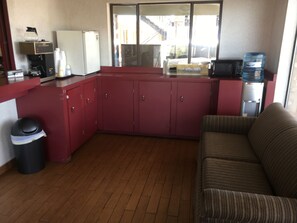 A compact kitchen with a microwave, refrigerator, and coffee maker. A water dispenser is on the counter, and a trash bin is visible.