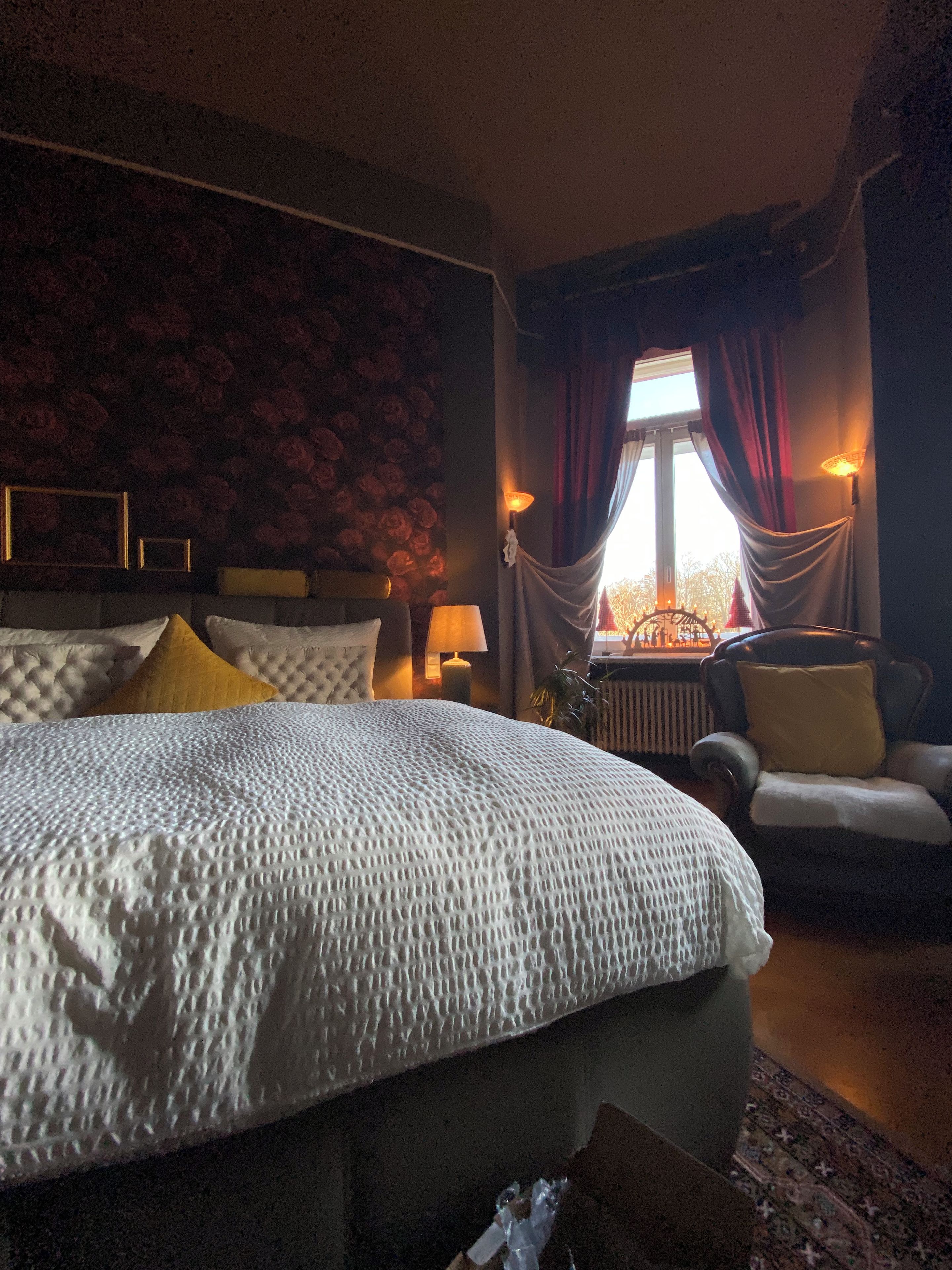 A hotel room with a large bed, a chair, a window with curtains, and a wall with a floral pattern.