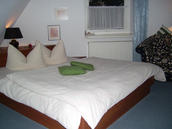Un dormitorio con cama, almohadas, una mesita de noche, lámpara, ventana con cortinas e imagen enmarcada en la pared.